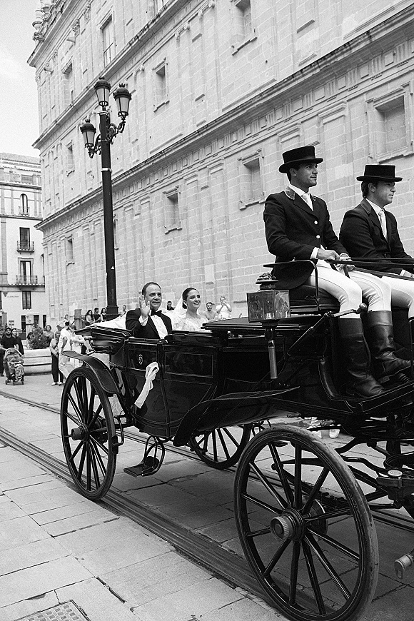 Wedding carriage exit as bride and groom ride in a horse-drawn carriage, waving to a crowd outside a historic stone building on a city street