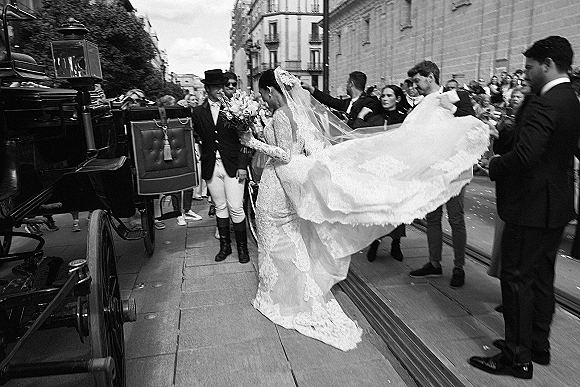 Wedding exit as bride steps into a horse-drawn carriage, lace gown train and veil gathered by groom in top hat on city street with guests