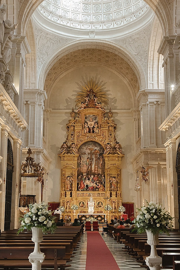 Church ceremony setup with red aisle runner, white urn floral arrangements, and candles leading to a gilded altar in an ornate cathedral interior