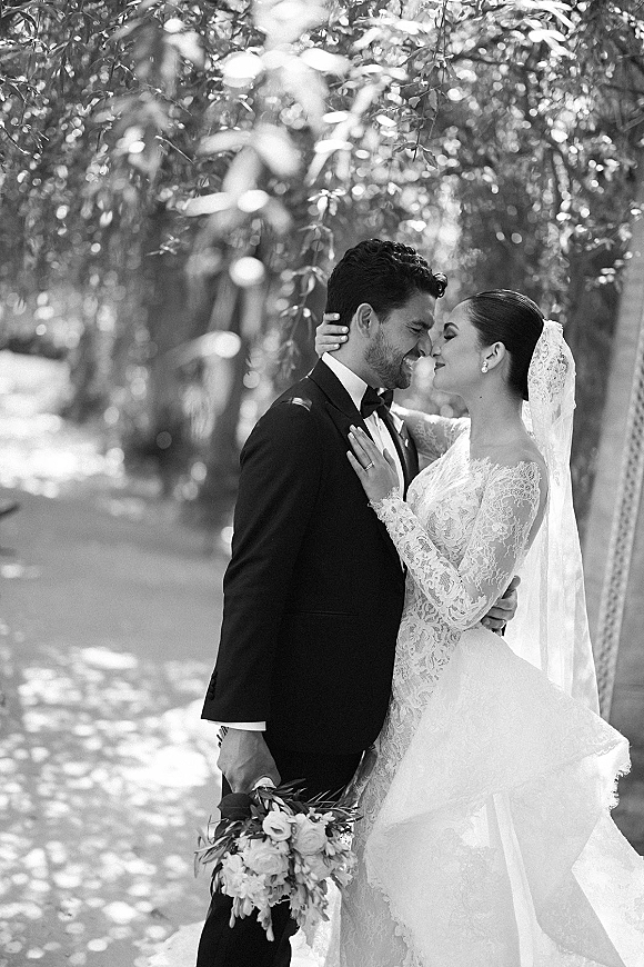 Wedding kiss portrait in black and white of bride and groom kissing on a tree-lined path, her veil and lace sleeves in dappled sunlight