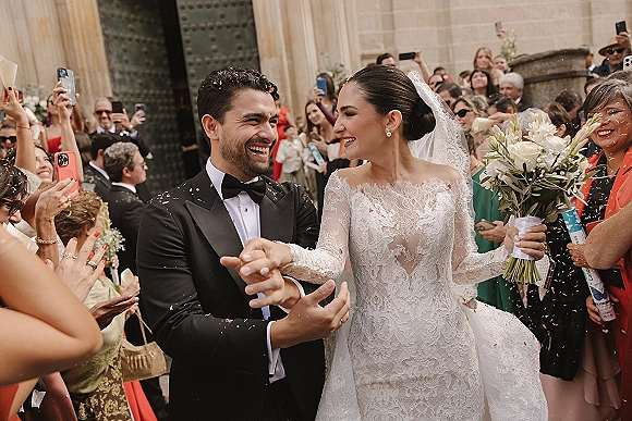 Wedding recessional as newlyweds walk out of a church entrance on stone steps, holding hands while guests toss confetti and film on phones
