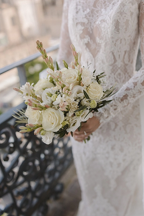 Bridal bouquet of white roses and greenery held against a lace wedding dress, engagement ring visible on an outdoor balcony railing