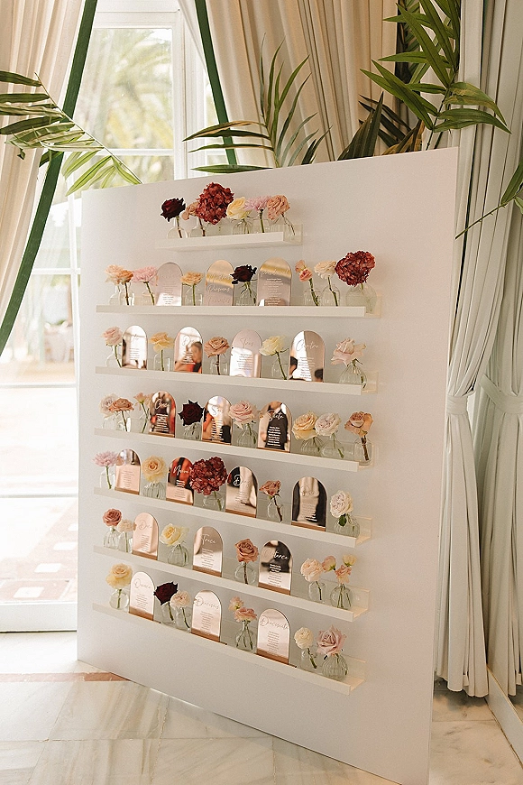 Wedding seating chart on a white display board with acrylic seating chart arch cards, bud vases, roses, and hydrangeas in window light