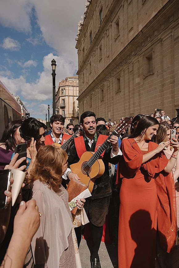 Wedding procession moving down a street as guests cheer and film, with an acoustic guitar player near a historic stone building backdrop