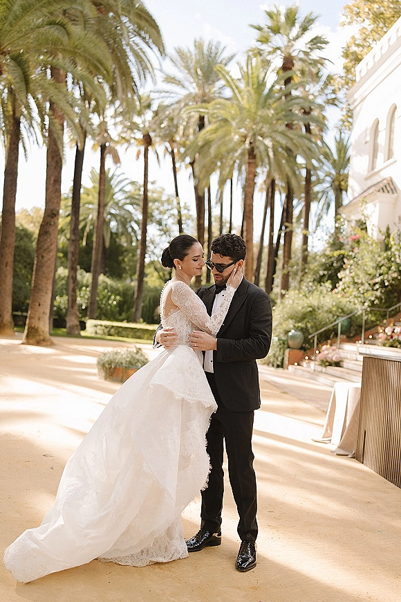Couple portrait of bride and groom embrace as she touches his face, her lace wedding dress train flowing on a sunlit palm-lined walkway