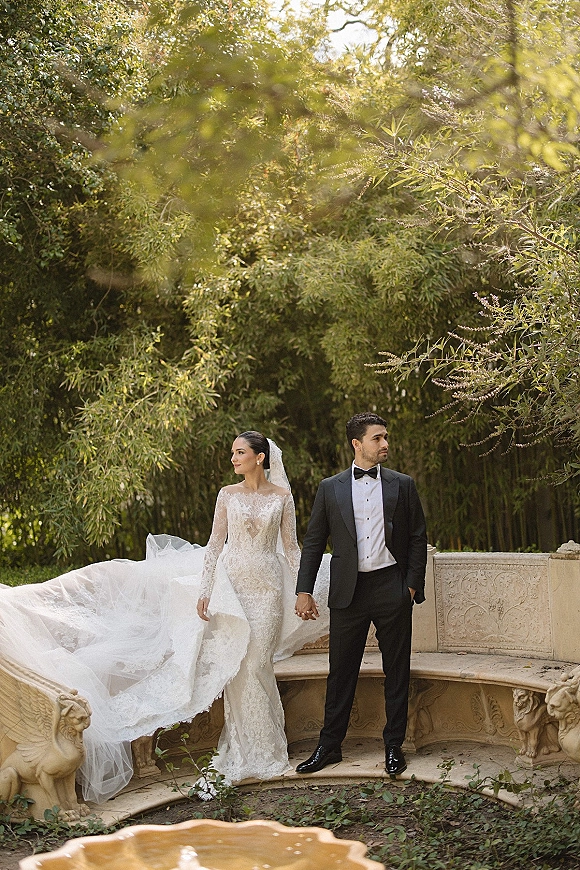 Couple portrait of bride and groom holding hands, bride in lace long sleeve dress and cathedral veil by a stone fountain in greenery