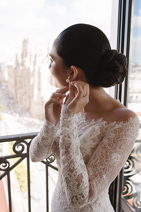 Bridal portrait of a bride putting on pearl drop earrings in window light, wearing a lace wedding dress on a balcony with city skyline