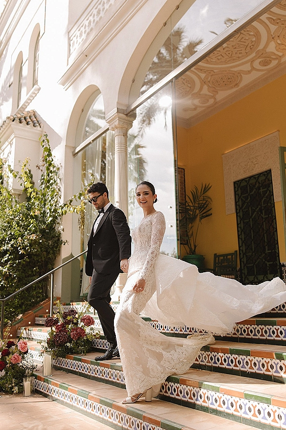 Couple portrait of bride and groom walking hand in hand down a tiled staircase, her lace long-sleeve dress train trailing by candles