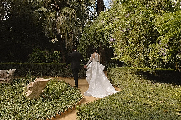 Couple portrait of bride and groom walking away holding hands, her open-back lace dress and train flowing on a garden path by statues