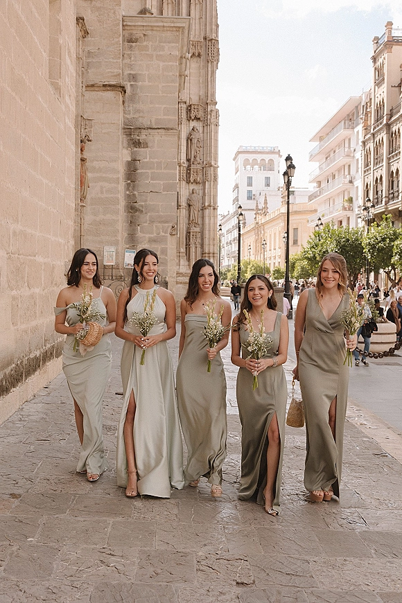 Bridesmaid group photo of women in sage green bridesmaid dresses walking with bouquets on a cobblestone street by a stone cathedral