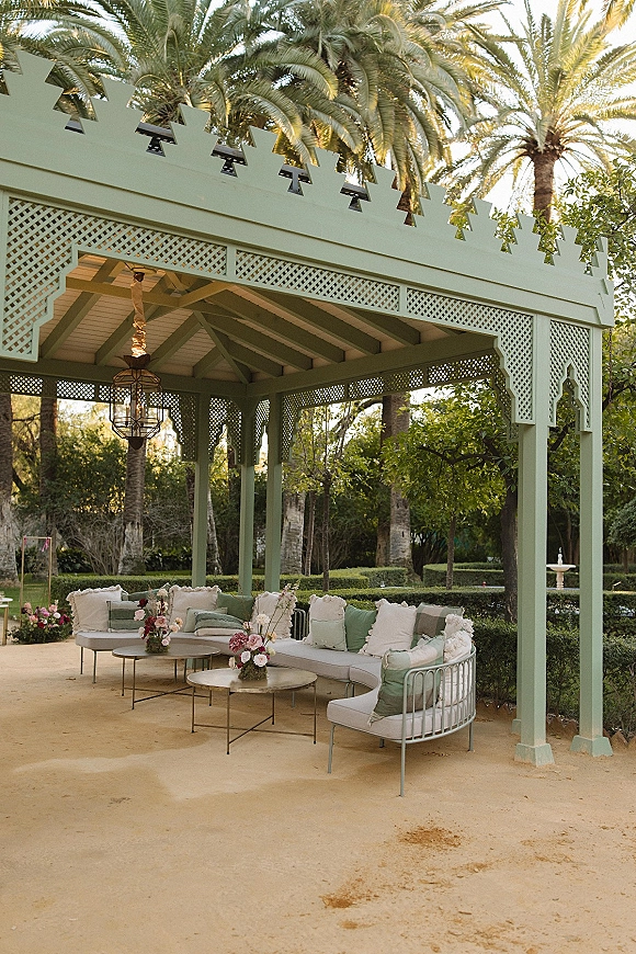 Outdoor lounge setup with a sectional sofa, throw pillows and floral centerpieces on coffee tables beneath a hanging lantern chandelier on a garden patio pergola