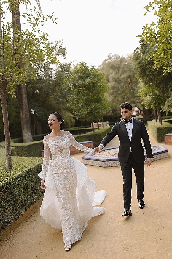Couple portrait of bride and groom walking hand in hand, her long sleeve lace dress with train flowing on a garden path by a tiled fountain