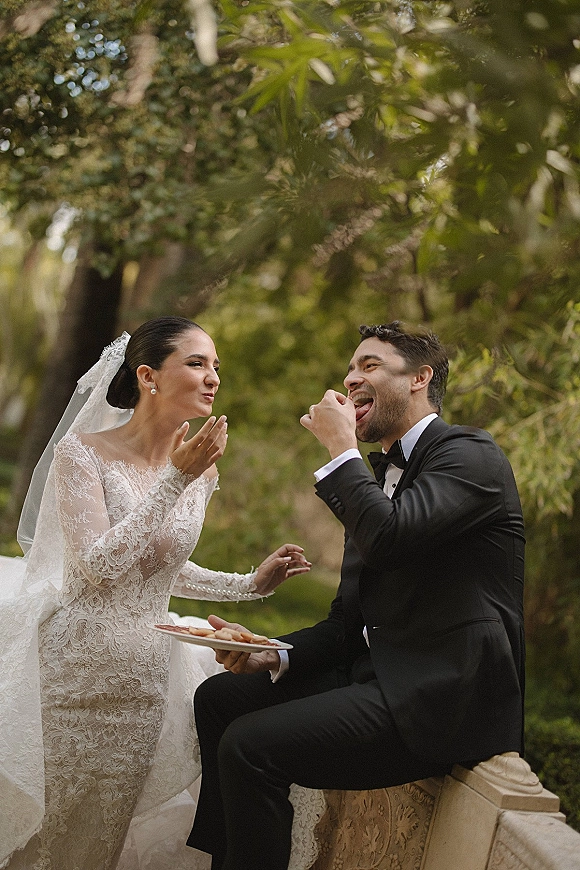 Couple portrait outdoors, bride in lace long sleeve wedding dress and veil laughing with groom as they share dessert by stone railing