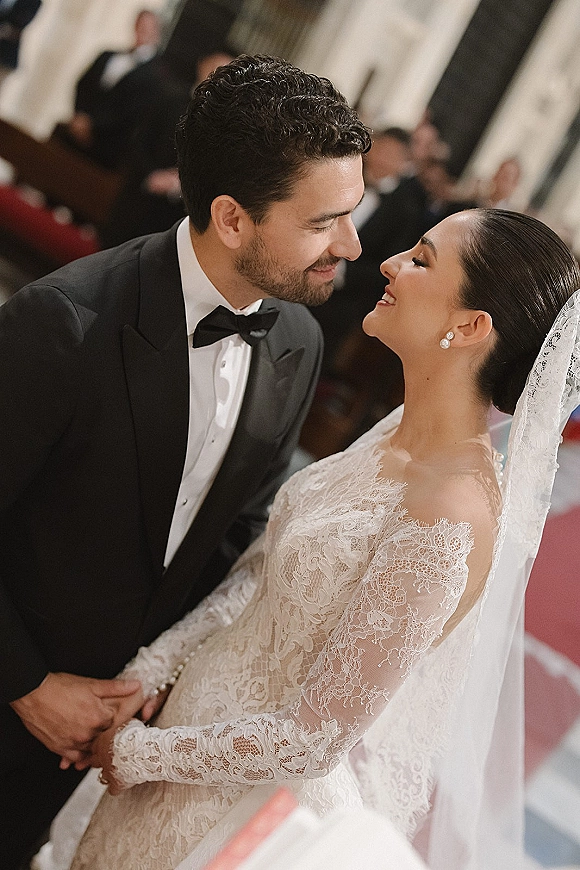 Wedding couple portrait of bride in lace wedding dress and groom in black tuxedo holding hands in a church aisle with guests behind