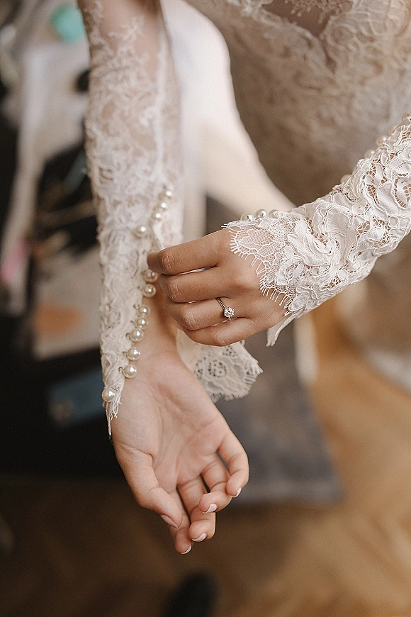 Wedding dress detail with lace sleeve cuffs and pearl buttons as the bride’s ringed hand adjusts them in a blurred getting-ready room