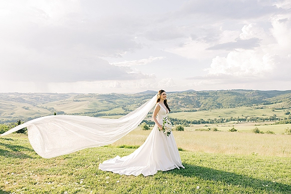 Bridal portrait of a bride holding bouquet, wearing a simple A-line wedding dress with long veil flowing over a grassy hillside under clouds