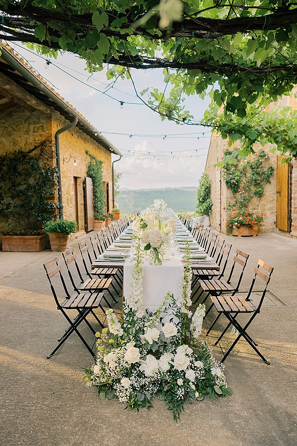Reception tablescape with a long banquet table wedding setup, white runner, rose and greenery centerpieces, candles, and string lights in a stone courtyard