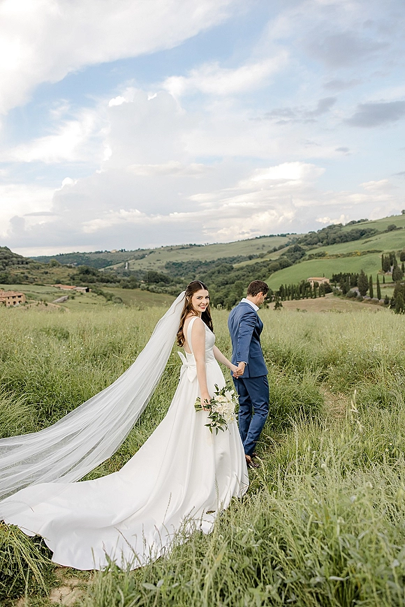 Couple portrait of bride and groom holding hands, bride looking back with long veil and bouquet on a dirt path through rolling hills