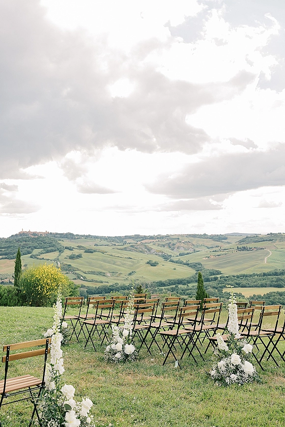 Outdoor ceremony setup with garden ceremony seating, wood folding chairs and white aisle florals on a grassy lawn with rolling hills beyond