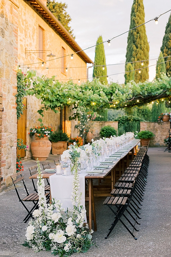 Outdoor reception tablescape with a long wooden banquet table, white florals and eucalyptus garland, candles, and string lights in a stone courtyard