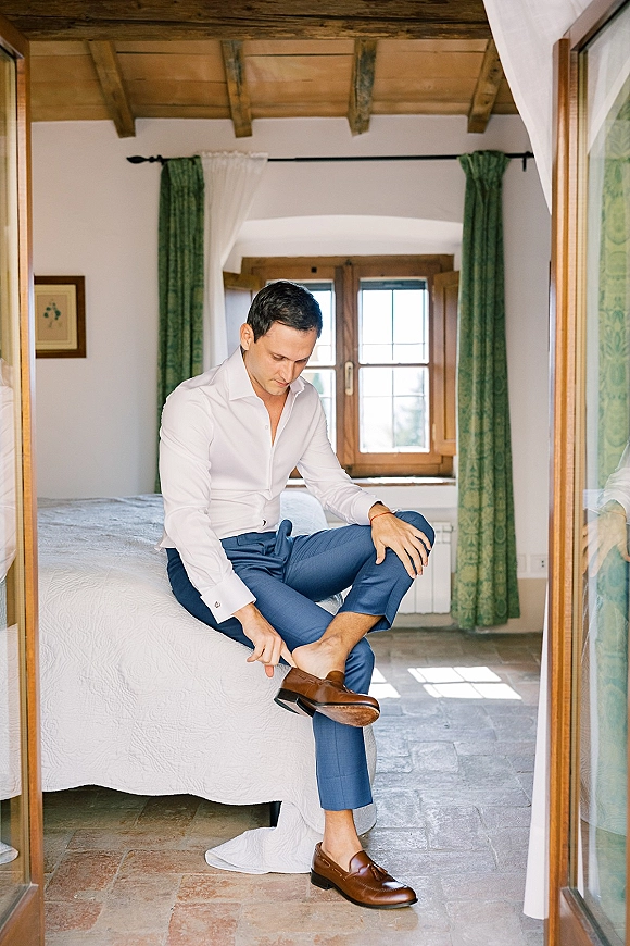 Groom getting ready, putting on brown leather loafers while sitting on the bed in a rustic bedroom with window light and green curtains