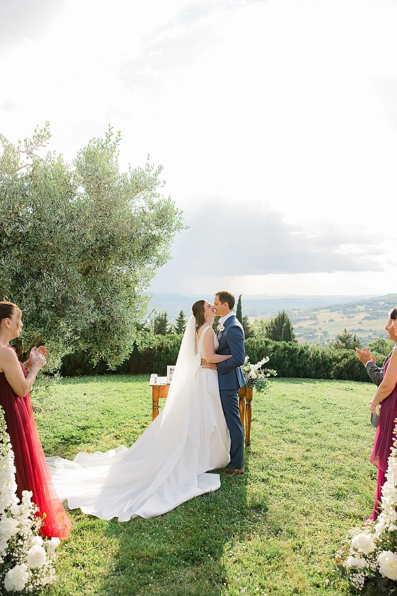 Ceremony kiss as bride in a wedding dress with long veil and train embraces groom in blue suit on a lawn with olive trees and hills
