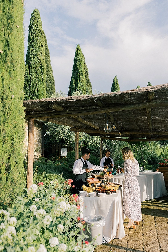 Outdoor cocktail hour buffet table with white linens, glassware, and bottled drinks under a wooden pergola with pendant lights in a garden
