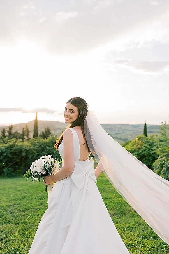 Bridal portrait of a bride looking back over her shoulder, long veil flowing, holding a bouquet on a lawn with sunset hills behind