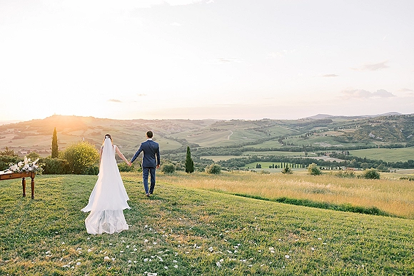 Couple portrait of bride and groom walking away holding hands, her long veil blowing at sunset over rolling hills and meadow grass