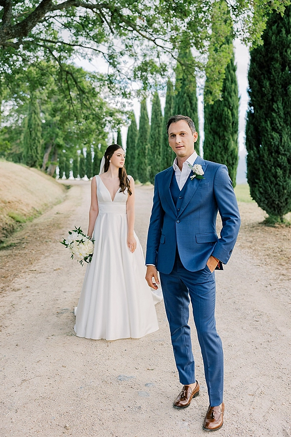 Couple portrait in an outdoor wedding portrait, bride in veil holding a white bouquet beside groom in a blue suit on a tree-lined gravel road