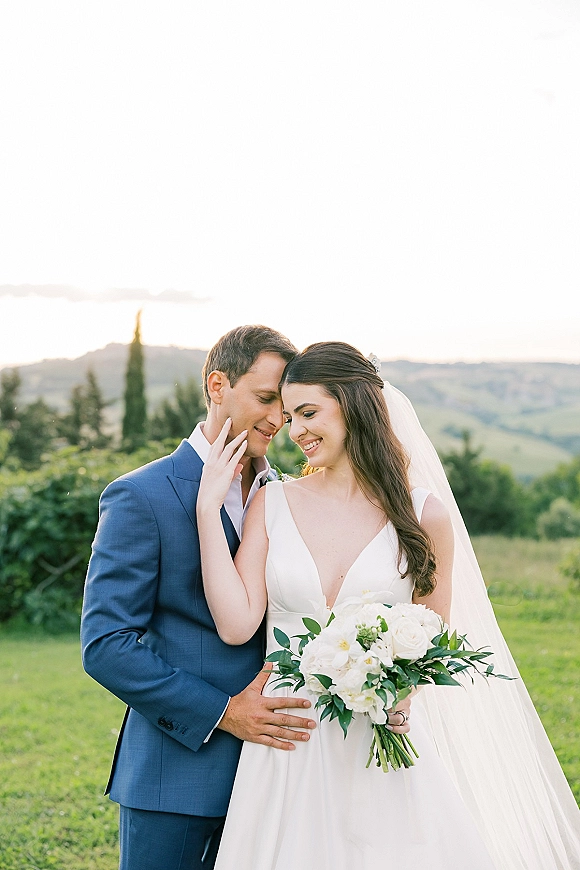 Couple portrait of bride and groom embracing, bride touching his face, holding a white bouquet in a grassy field with rolling hills behind
