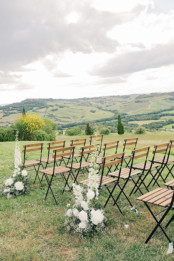 Ceremony aisle decor with white rose and baby's breath aisle markers lining wood folding chairs in a grassy field with rolling hills under clouds
