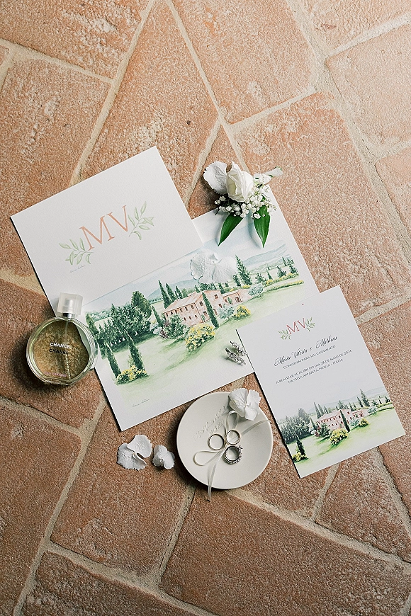 Invitation suite wedding invitation flat lay with watercolor cards, monogram, rings in dish, perfume, ribbon, petals on terracotta tile floor