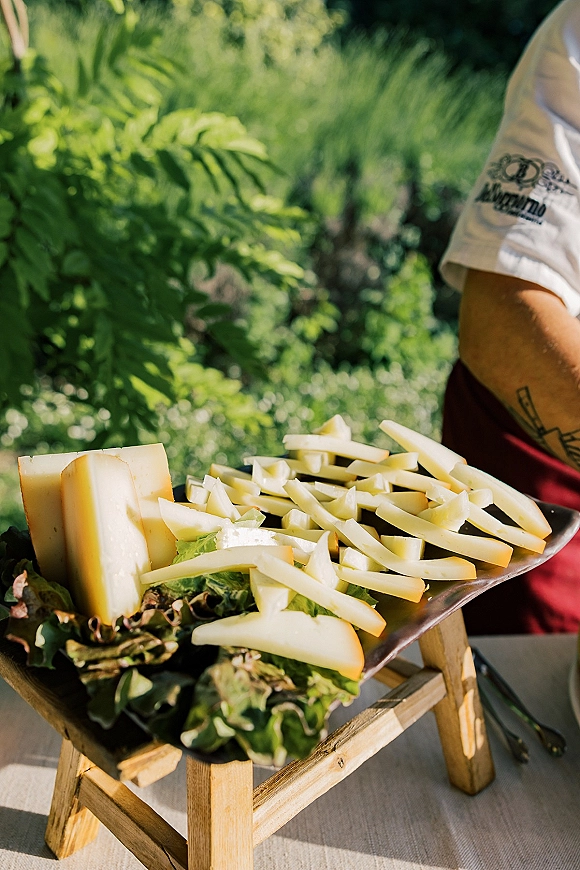 Cheese platter with sliced cheese and wedges on a wooden serving stand, accented by leafy greens on a sunlit garden tablecloth
