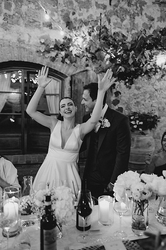 Reception couple moment as newlyweds at dinner table, bride in wedding dress raising arms while groom smiles under string lights by stone wall