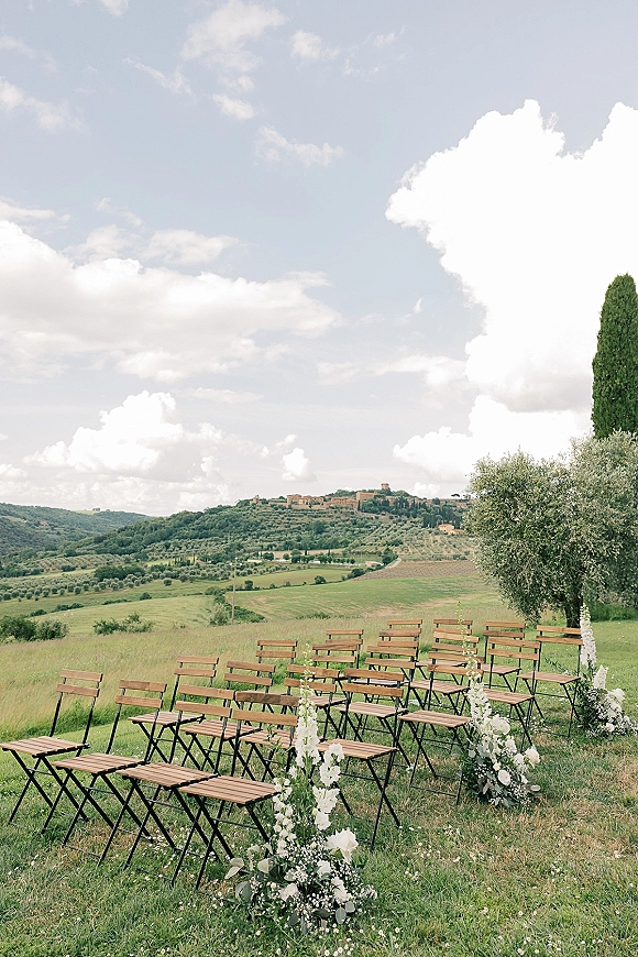 Outdoor ceremony setup with wood folding chairs and white floral aisle arrangements on a grassy field, rolling hills and cloudy sky beyond