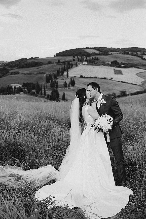 Wedding kiss as bride and groom embrace on a grassy hillside, her veil and bouquet visible against rolling farmland hills under cloudy sky