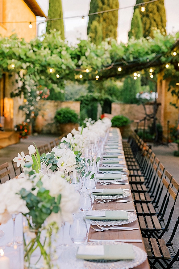 Reception tablescape with long banquet wedding table, white florals and eucalyptus garland, taper candles and string lights in a courtyard