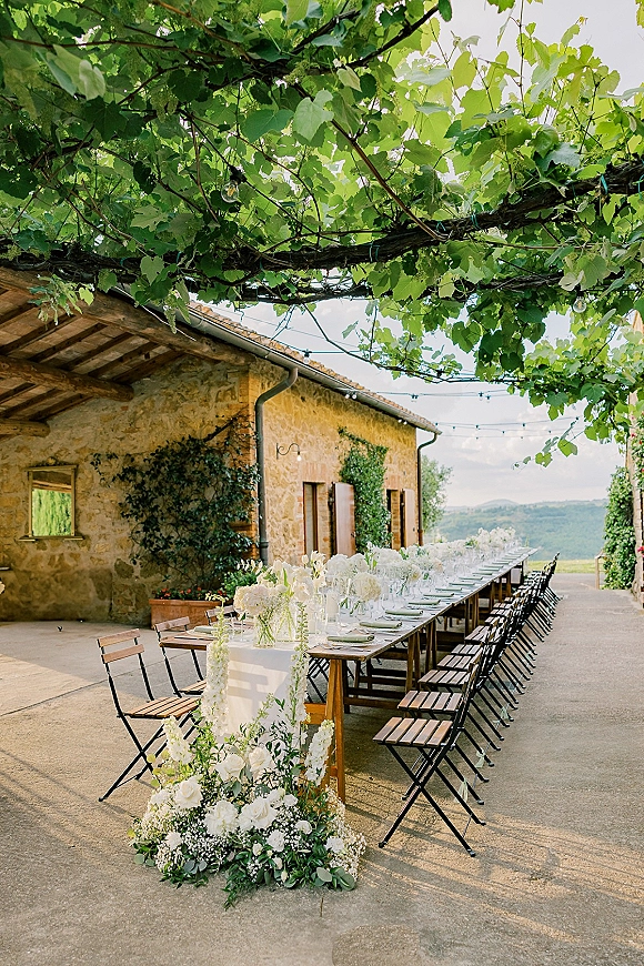 Outdoor reception tablescape with a long banquet table wedding setup, white runner, florals and greenery under string lights by a stone farmhouse wall
