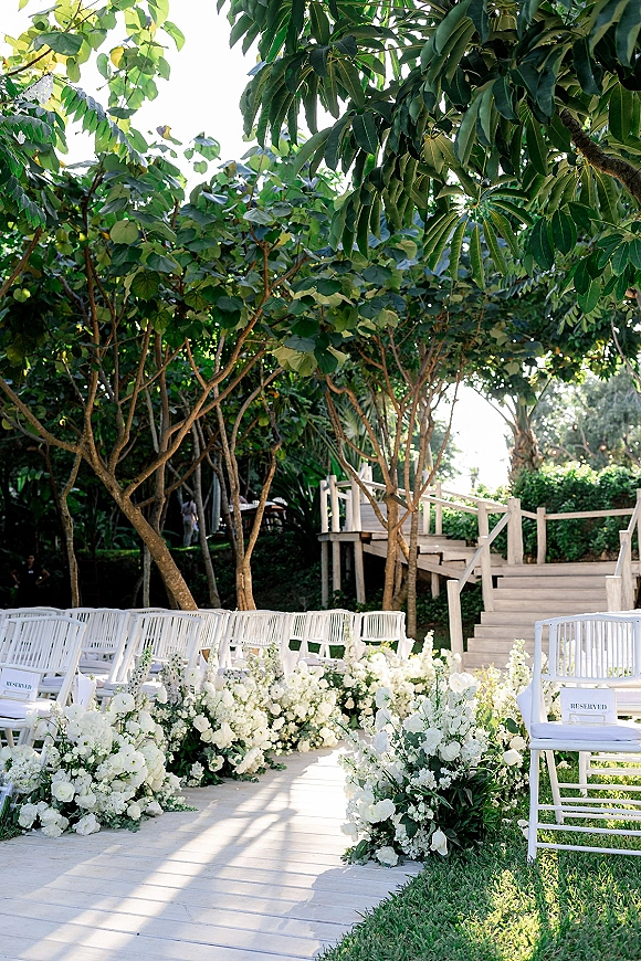 Ceremony aisle decor with white florals and greenery lining a wood plank aisle, reserved white chairs and railing in a sunlit garden setting
