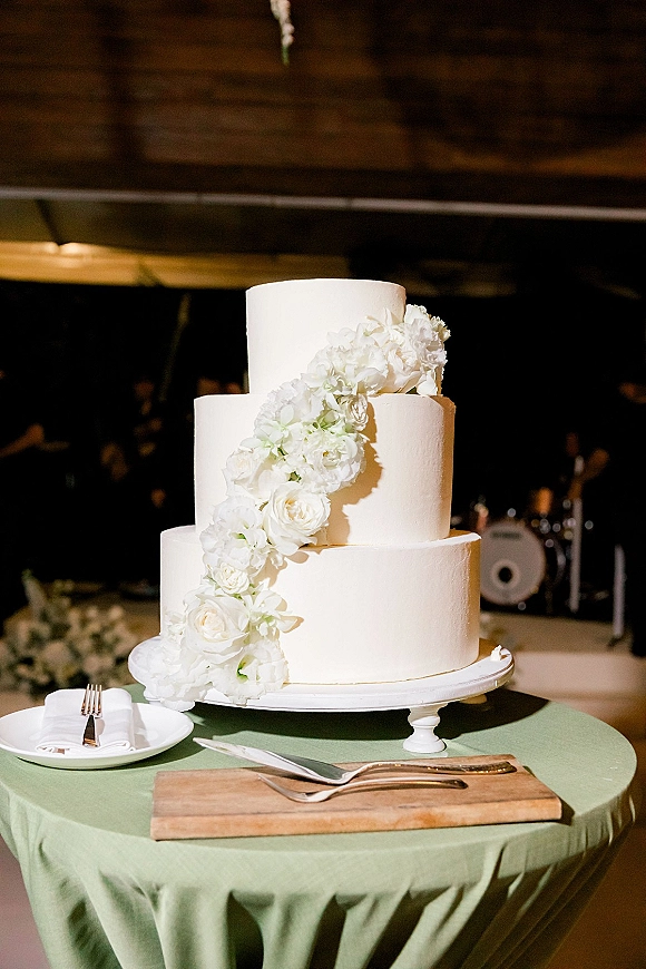 Wedding cake with a white floral cascade on a stand, set on a green tablecloth with knife and server in a dim reception space