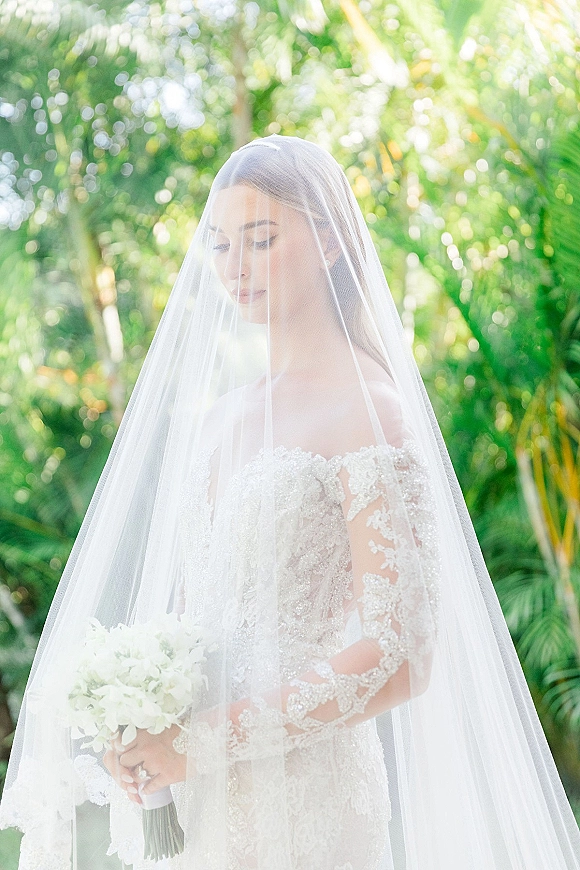 Bridal portrait of a bride with veil in a lace gown, holding a white bouquet, with palm trees and tropical garden greenery behind her.