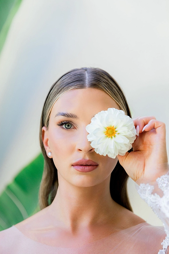 Bridal portrait of a bride holding flower over one eye, wearing pearl studs and lace sleeve against a neutral wall with a green leaf