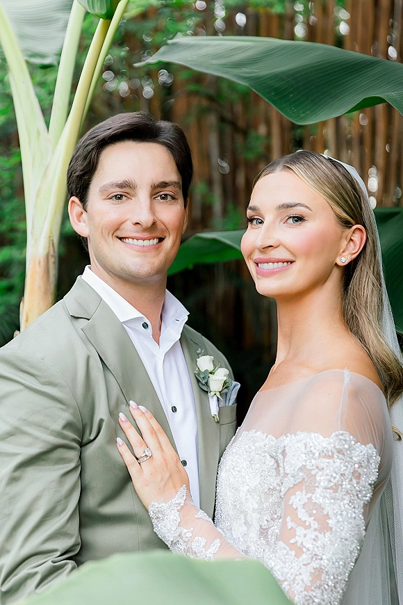 Couple portrait of bride and groom close up, bride’s veiled hand with ring on his chest, framed by tropical leaves and wooden fence