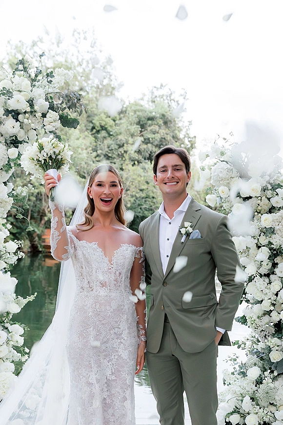 Couple portrait of bride in lace veil holding a white bouquet with groom in sage suit beneath a white floral arch by a lakeside tree line