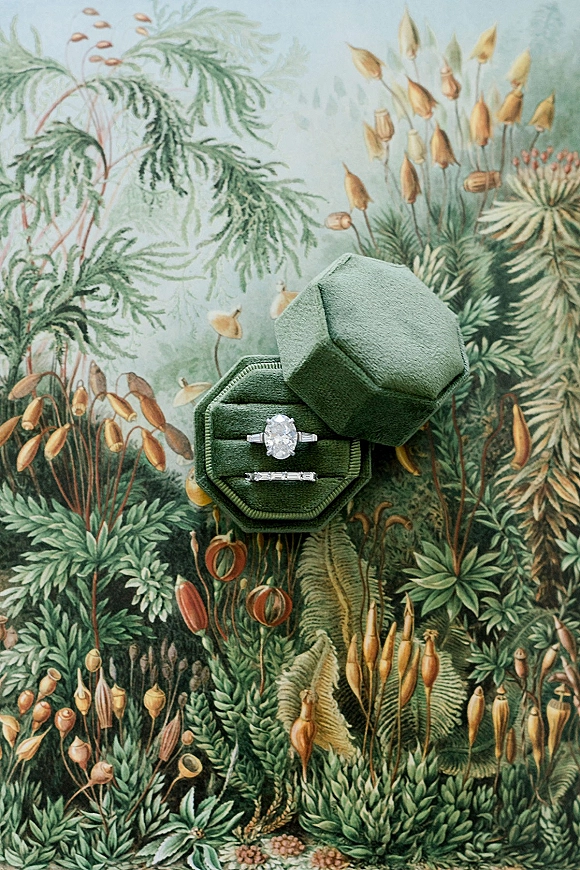 Engagement ring with an oval diamond ring stacked beside a wedding band in a velvet ring box on botanical wallpaper backdrop
