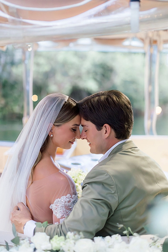 Wedding couple portrait with a forehead touch, bride in veil and lace off-shoulder dress holding white bouquet under clear tent lights