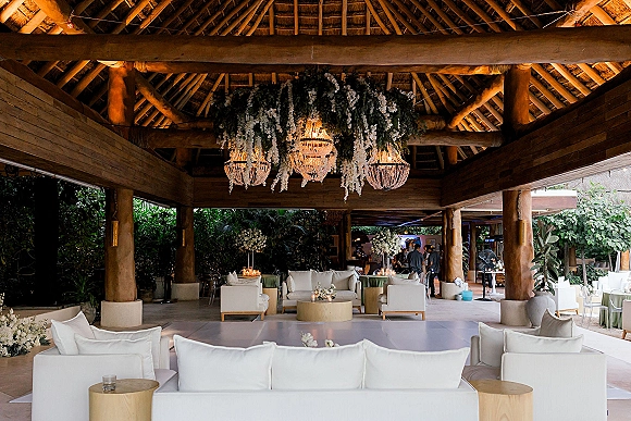 Reception lounge setup with white sofas and candlelit coffee tables beneath chandeliers and hanging florals in an open-air wood pavilion