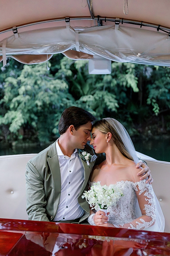 Couple portrait of bride and groom on boat, embracing with a white bouquet and veil, lake water and trees behind them