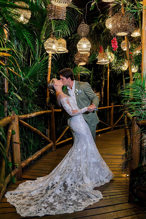 Wedding kiss portrait of bride and groom kiss in a dip on a lantern-lit wooden walkway, her lace long-sleeve train flowing at night